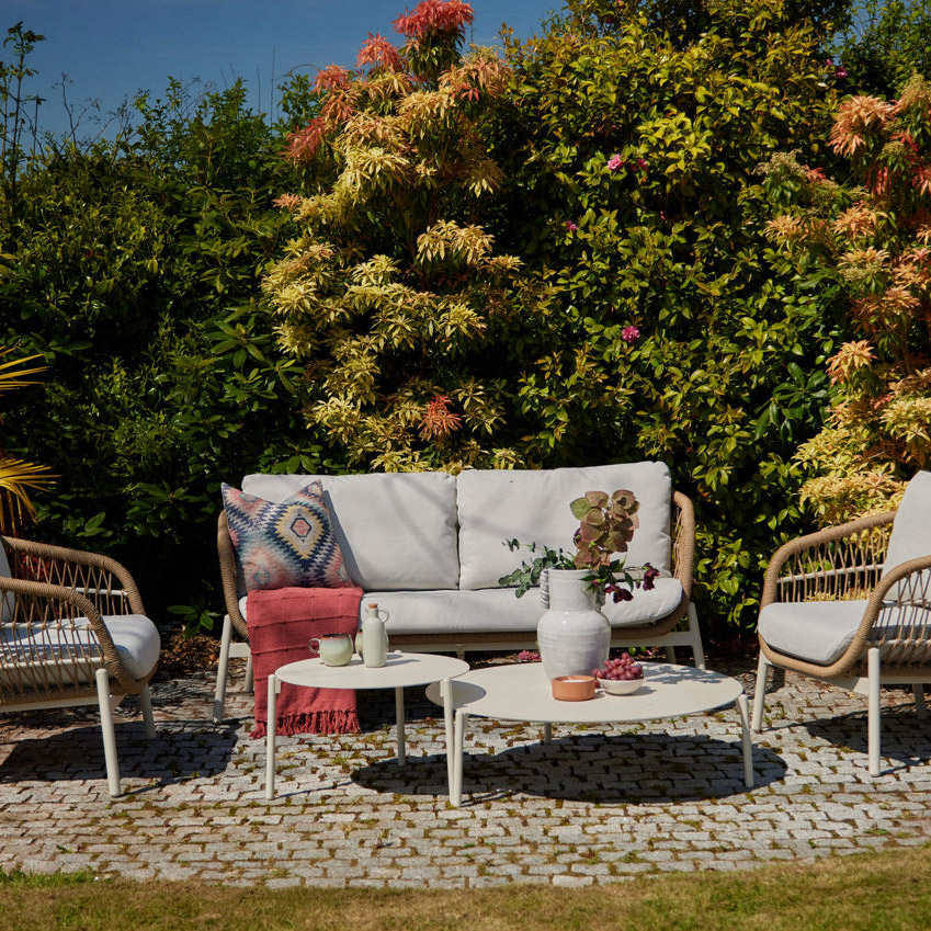 Outdoor seating set with four wicker chairs, a glass table, and tableware on a patio, surrounded by greenery.