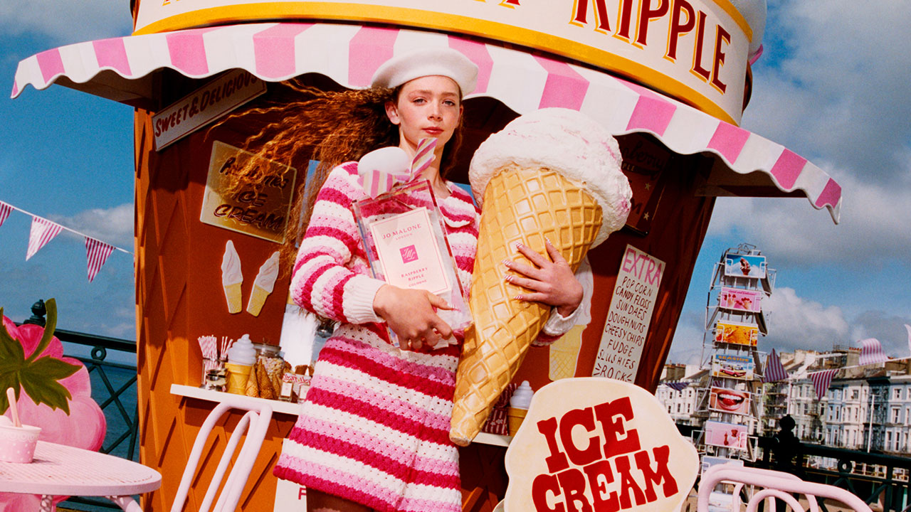 Model standing in a carnival holding an oversized prop of Jo Malone London Raspberry Ripple fragrance bottle and a giant ice cream cone