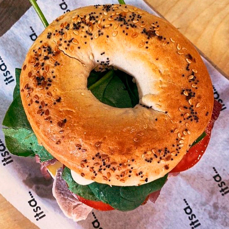 A seeded bagel sandwich with spinach, tomato, and meat on paper-lined surface.