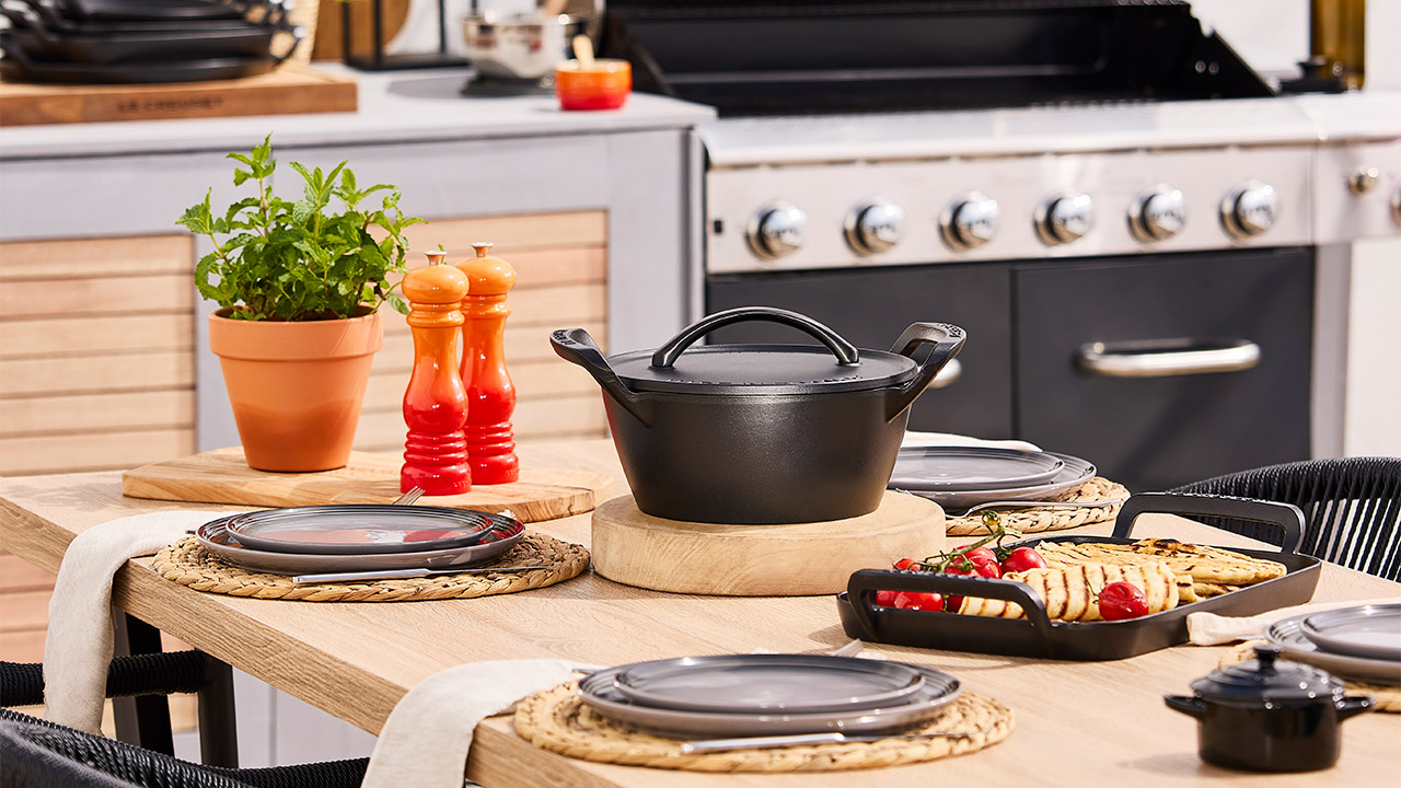 A dining table set with black dishes, a pot, a plant, and condiments, located in a kitchen with a stove in the background.
