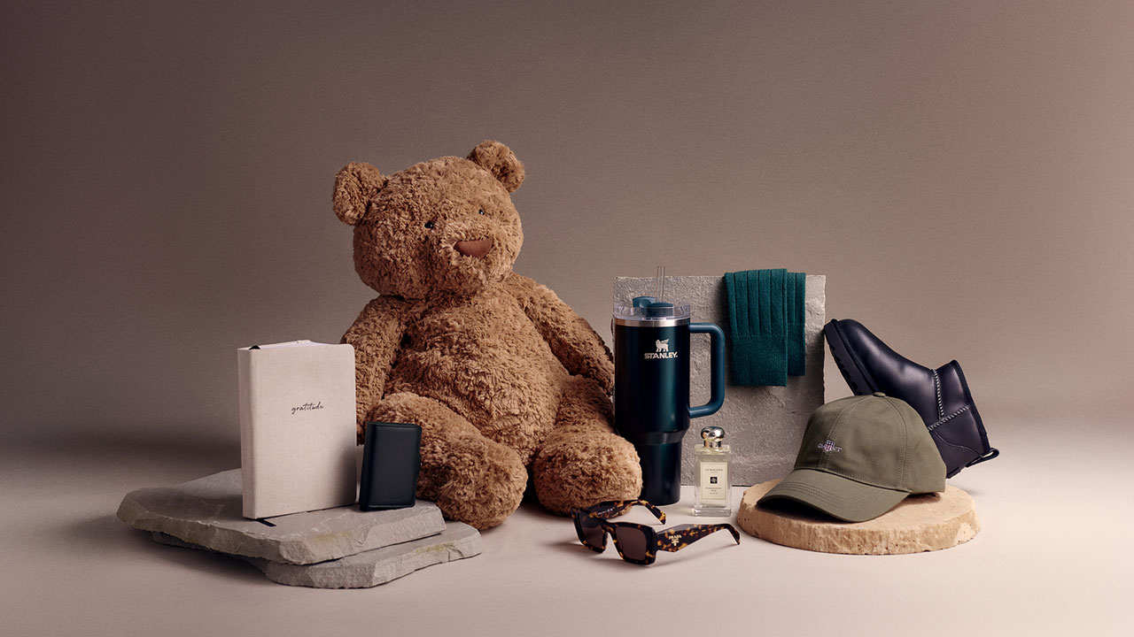A teddy bear is surrounded by books, a mug, sunglasses, a cap, shoes, a scarf, and a perfume bottle.