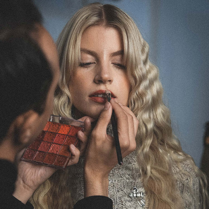 A person applying lipstick to a woman with long, wavy blonde hair, holding a palette of lip colors.