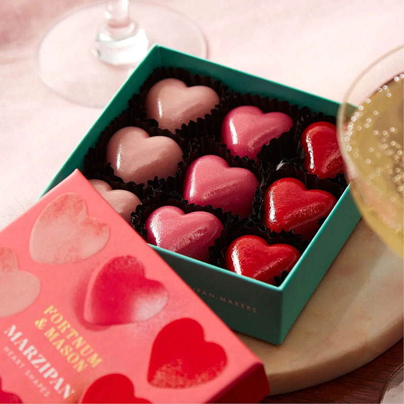 Heart-shaped chocolates in a teal box with a pink lid, alongside glasses with drinks.