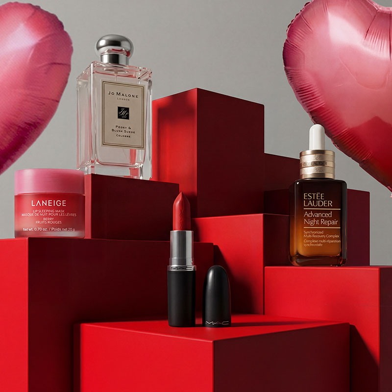 Cosmetics and perfume displayed on red cubes, surrounded by heart-shaped balloons.