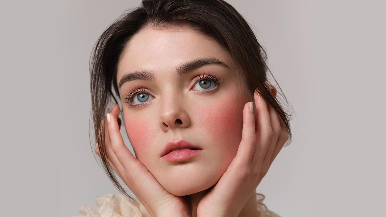 A woman with soft makeup and subtle blush, embodying bridal beauty, poses with hands gently cradling her face against a neutral background.

