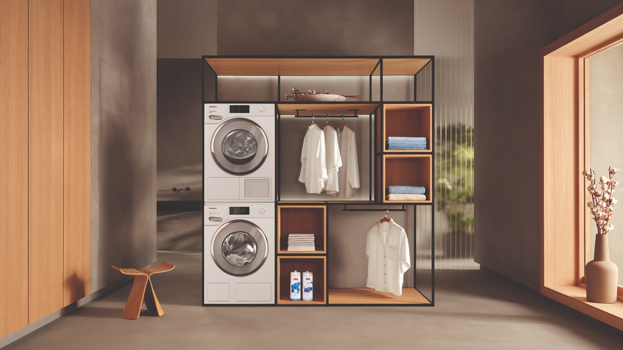 A modern laundry setup with Miele washer and dryer, clothes hanging, shelves with folded towels, and a minimalist decor.
