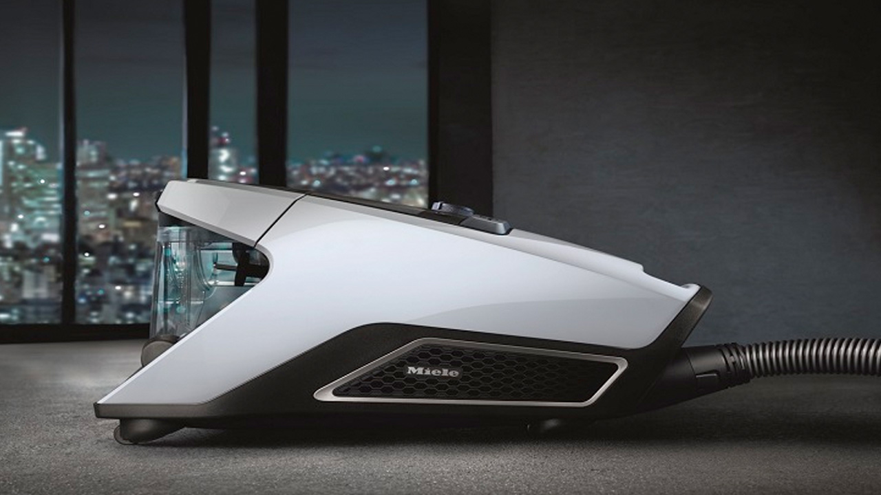 A sleek, modern Miele vacuum cleaner is placed in a room, with a cityscape visible through large windows in the background.