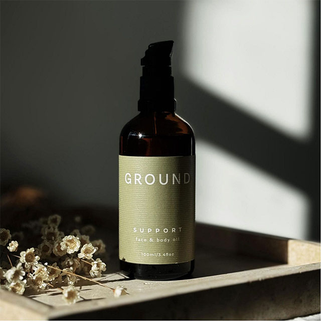 A bottle of 'Ground Support' face and body oil sits in sunlight, next to dried flowers.