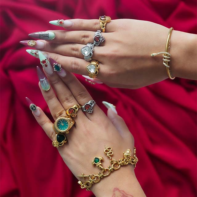 Hands with colorful, jeweled rings and bracelets over a red fabric background.