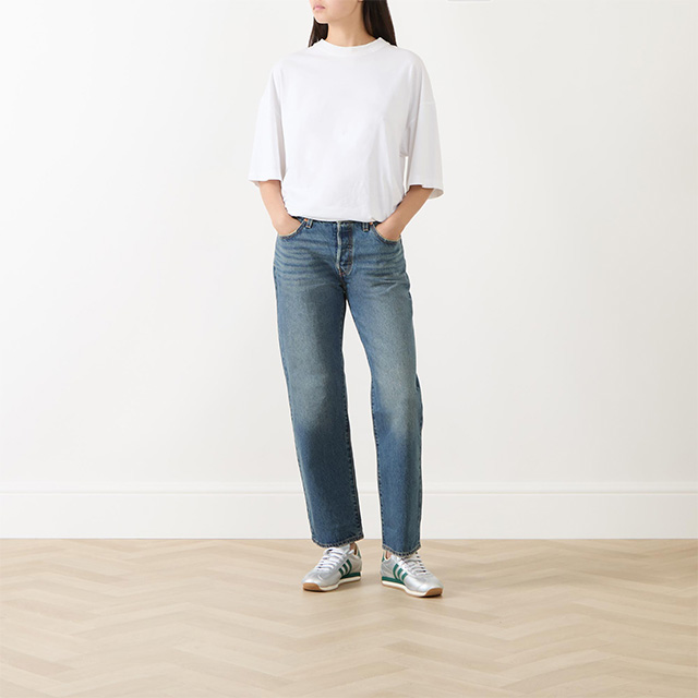 Person wearing a white t-shirt, blue jeans, and sneakers, standing on a wooden floor against a plain wall.