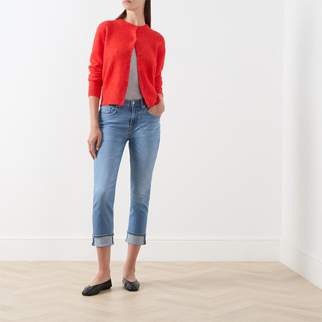 A person wearing a red cardigan, gray shirt, blue jeans, and black flats stands in a minimalistic room.