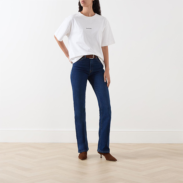 Person wearing a white t-shirt, blue jeans, and brown shoes stands in a minimalist room.