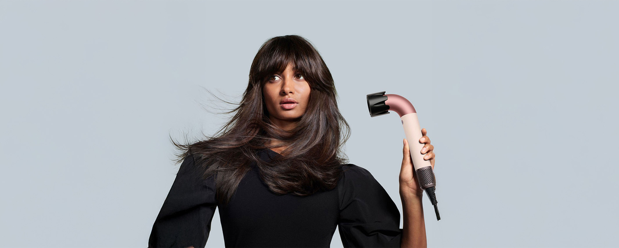 Person holding a hair styling tool with flowing hair, wearing a black top against a gray background.