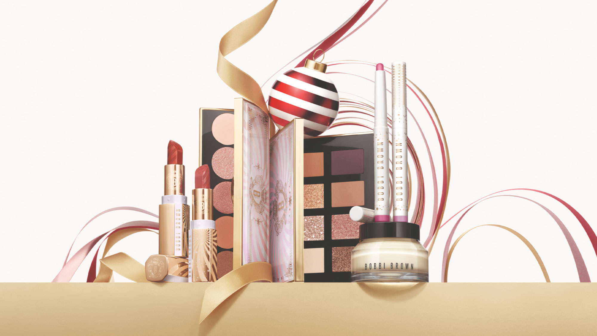 Makeup products and palettes with festive ribbons and a bauble, set against a light background.'
