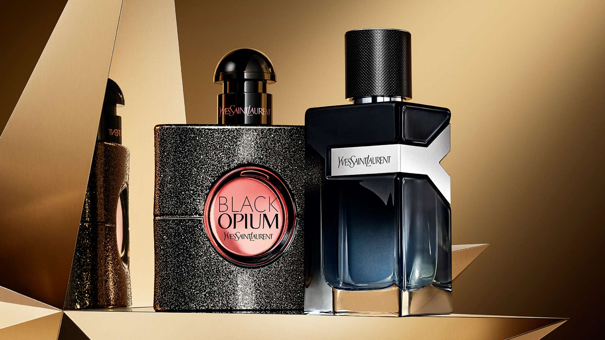 Two YSL perfume bottles, one black and glittery, the other dark blue, set against a geometric, gold-toned background.