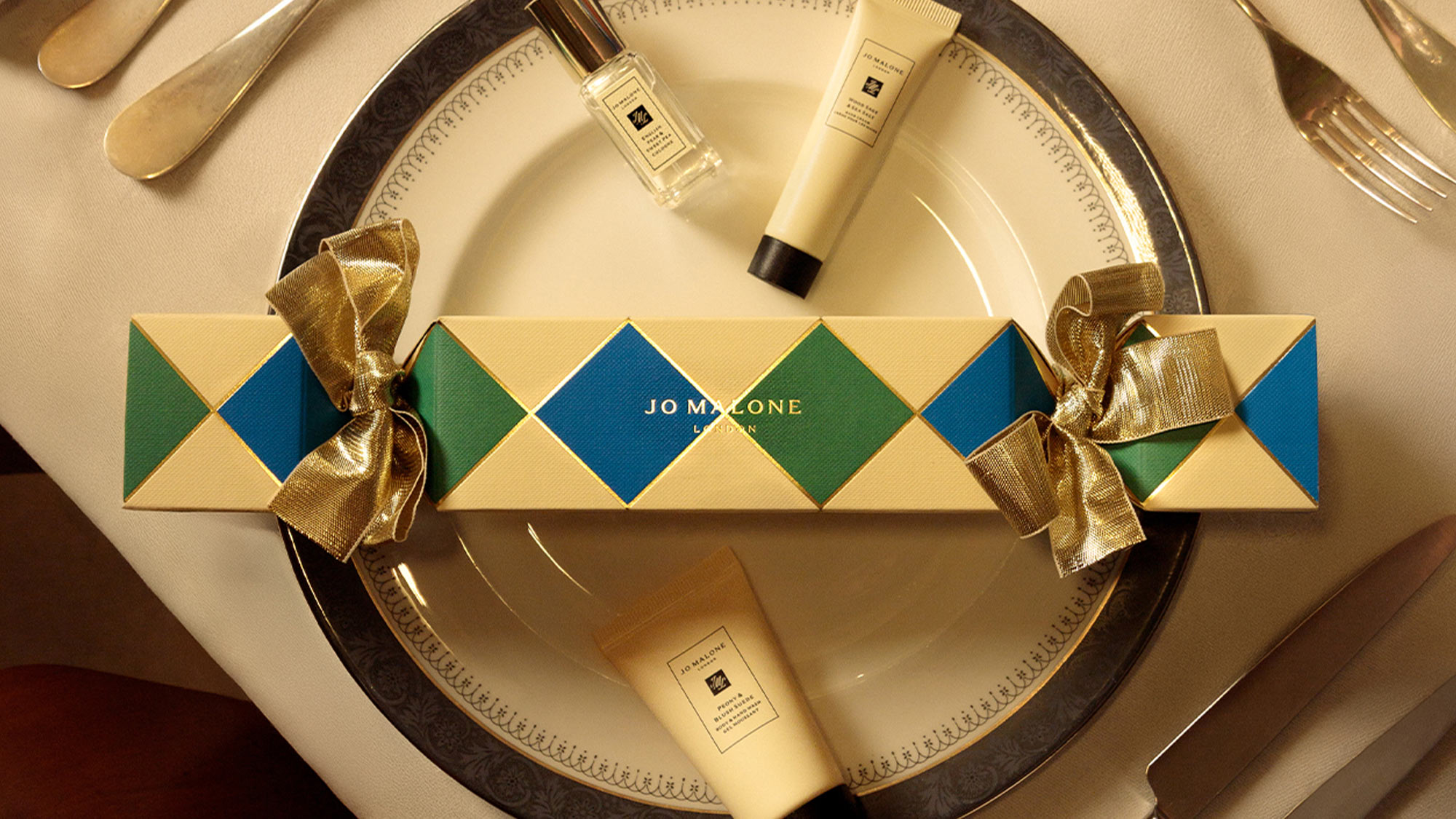 A gift box with blue and green diamond pattern, gold bows, and Jo Malone products on a decorative plate, surrounded by cutlery.