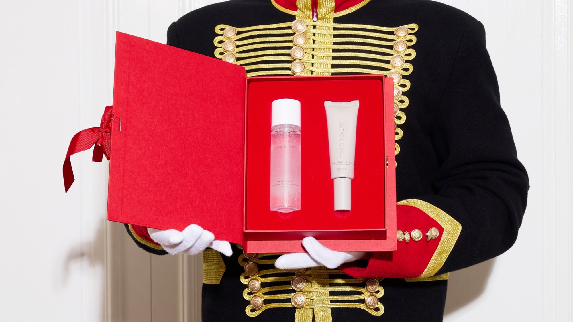 A person in a ceremonial uniform holds an open red box with two cosmetic products inside.