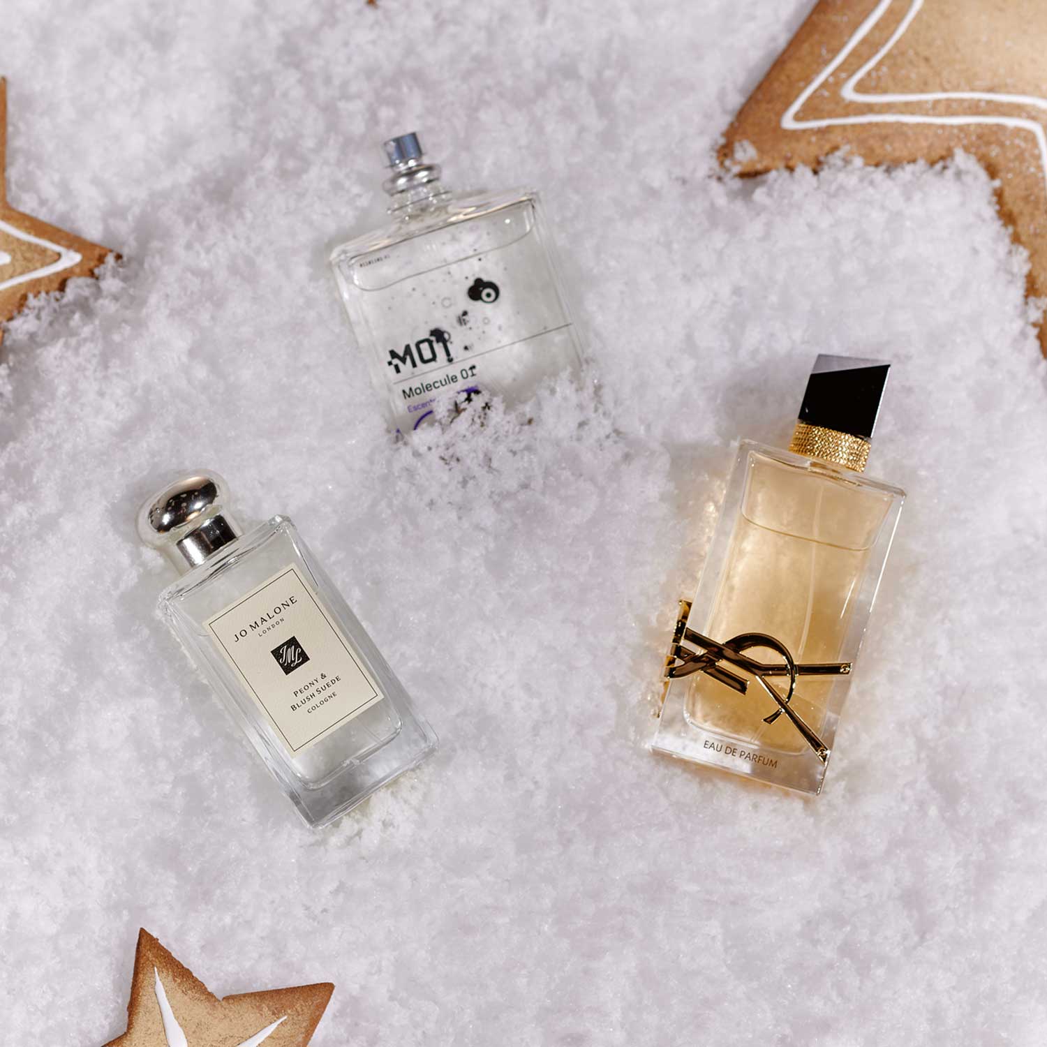 Three perfume bottles are arranged on snowy ground, surrounded by star-shaped gingerbread cookies.