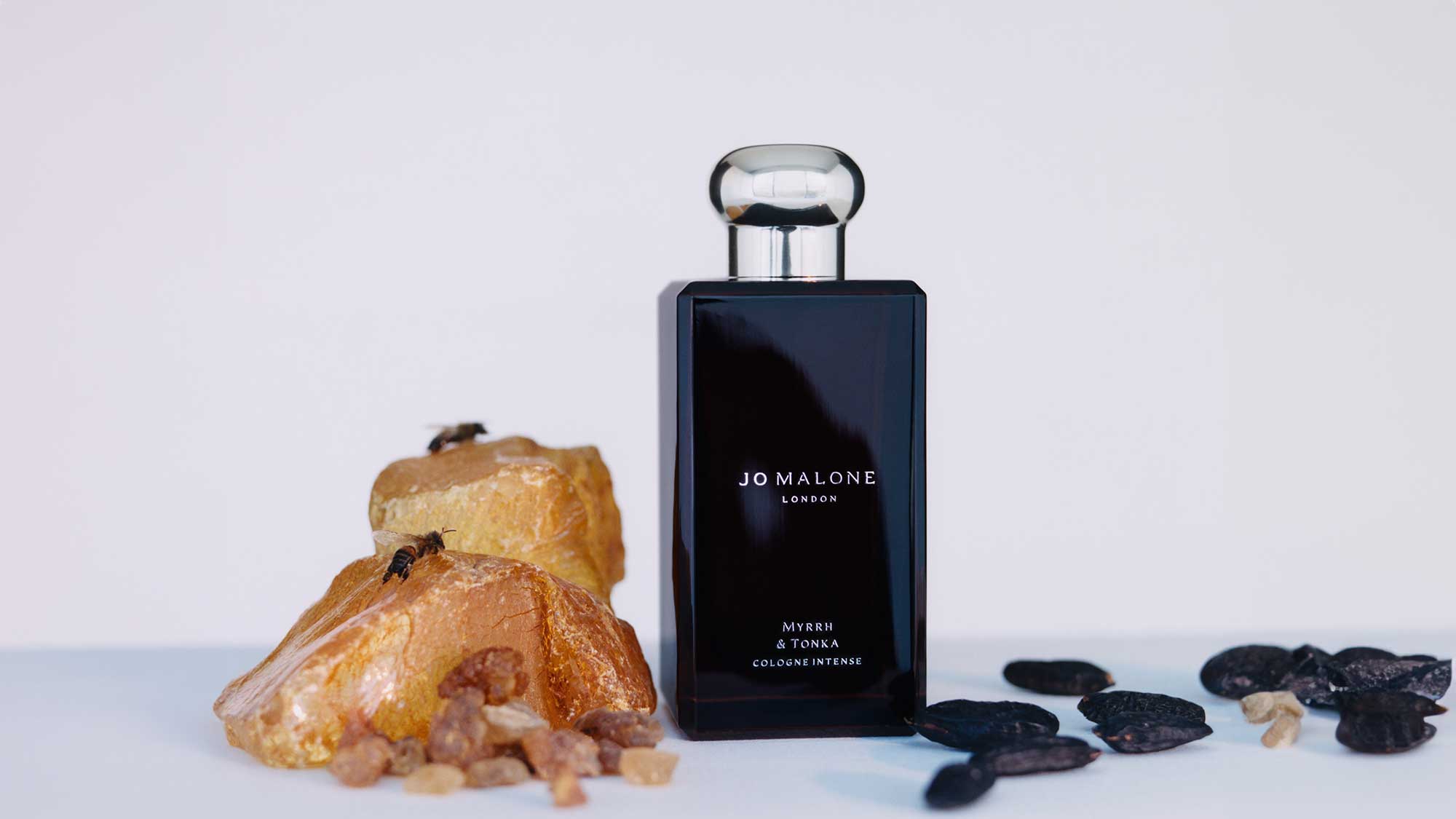A perfume bottle labeled 'Jo Malone London, Myrrh & Tonka, Cologne Intense' is placed beside amber-colored stones and black resin.