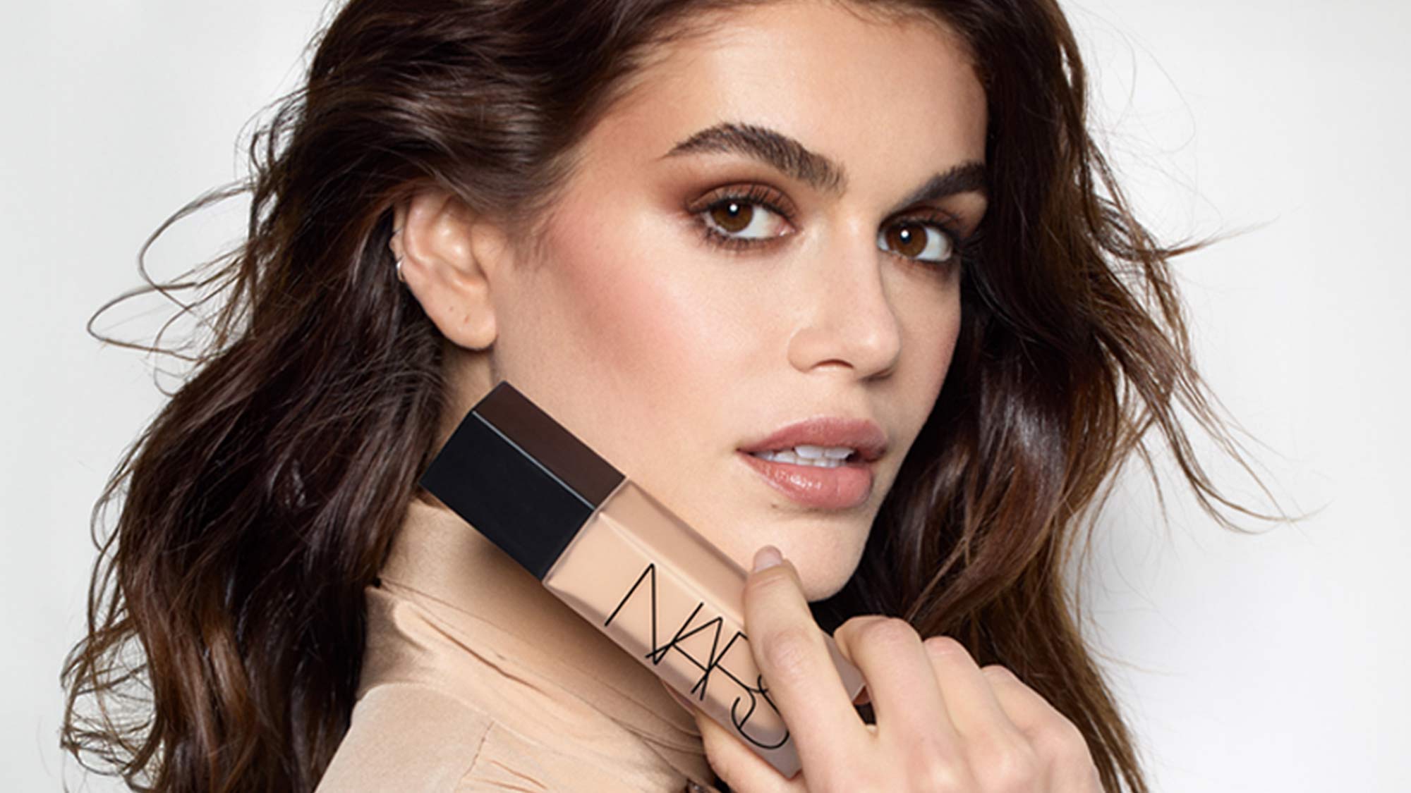 A person holds a NARS foundation bottle, showcasing makeup on their face with clean, soft brown hues.