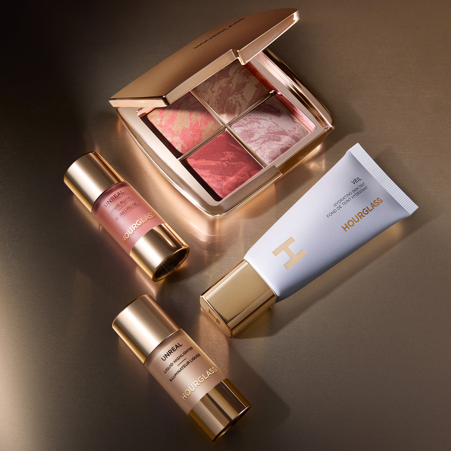 Luxury makeup collection featuring pink eyeshadow palette, rose-toned liquid highlighter, and primer bottles arranged on neutral background.