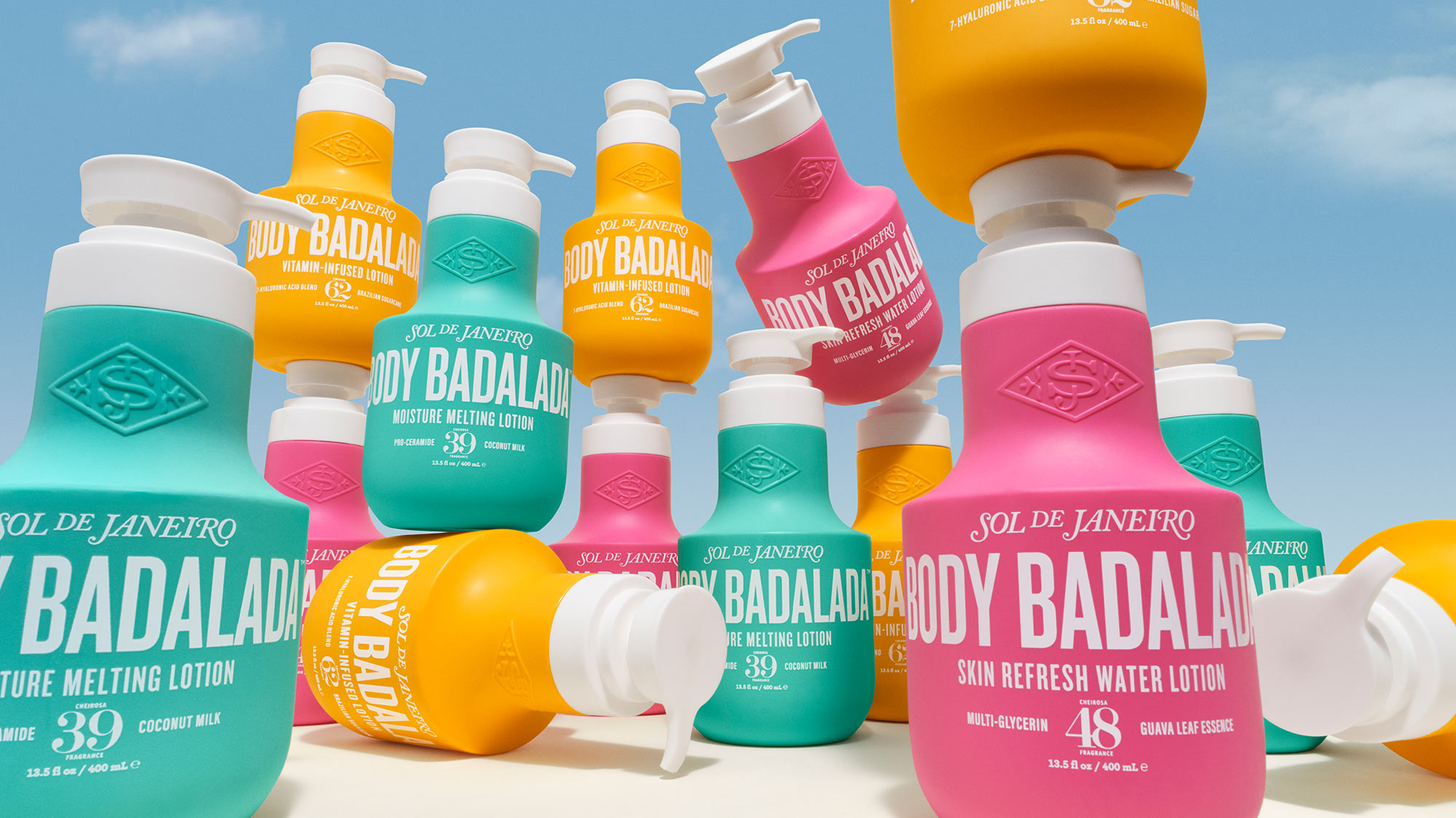 Collection of Sol de Janeiro Body Badalada lotions in turquoise, pink, and golden yellow bottles with pump dispensers against blue sky.