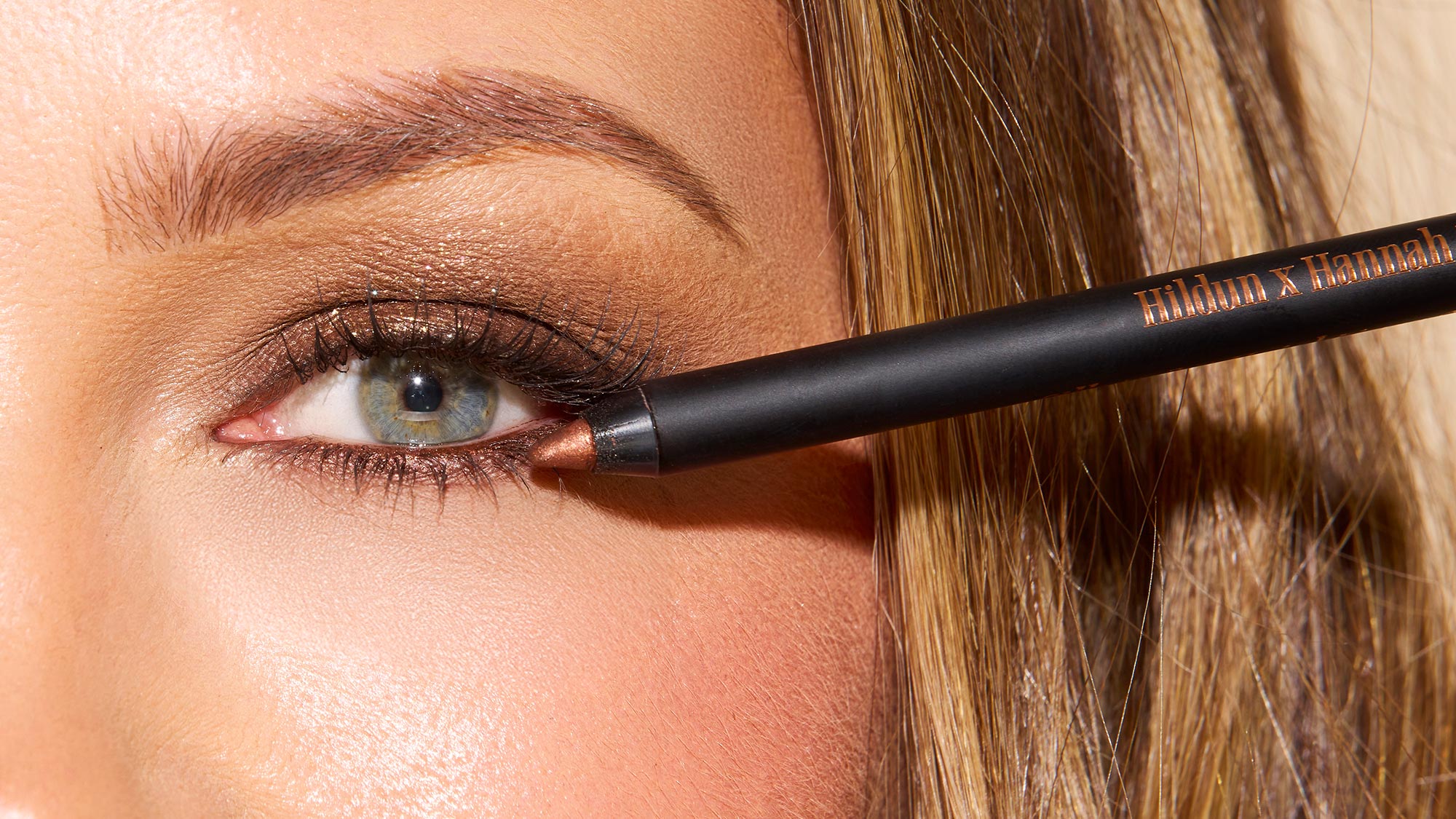 Close-up of person applying black eyeliner pencil to upper lash line with brown eyeshadow