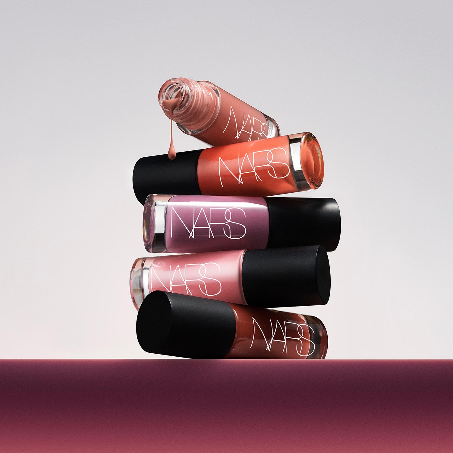 Stack of NARS Insatiable Liquid Blush bottles in coral, pink and burgundy shades with black caps, one dripping product visible