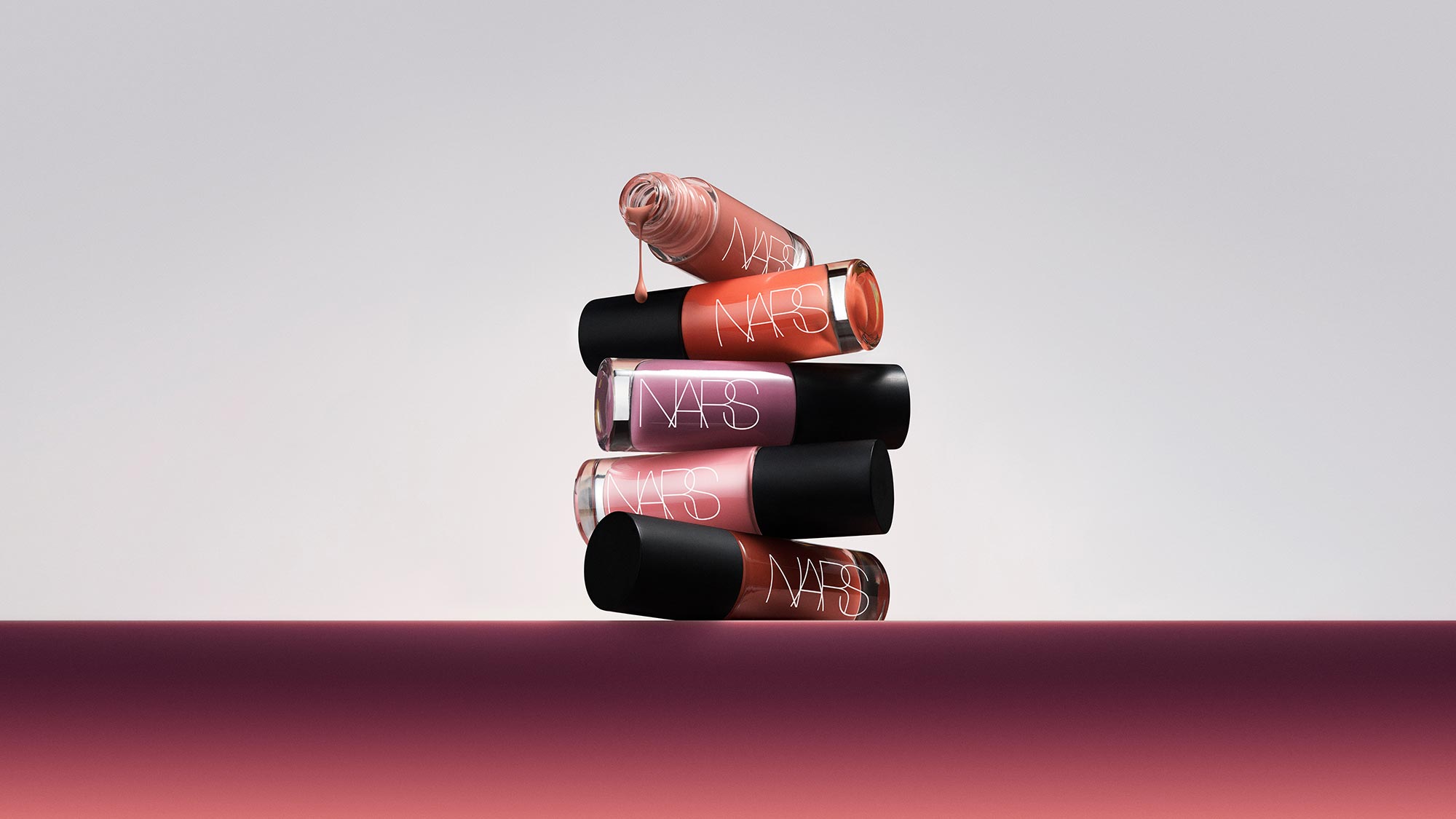 Stack of NARS Insatiable Liquid Blush bottles in coral, pink and burgundy shades with black caps, one dripping product visible
