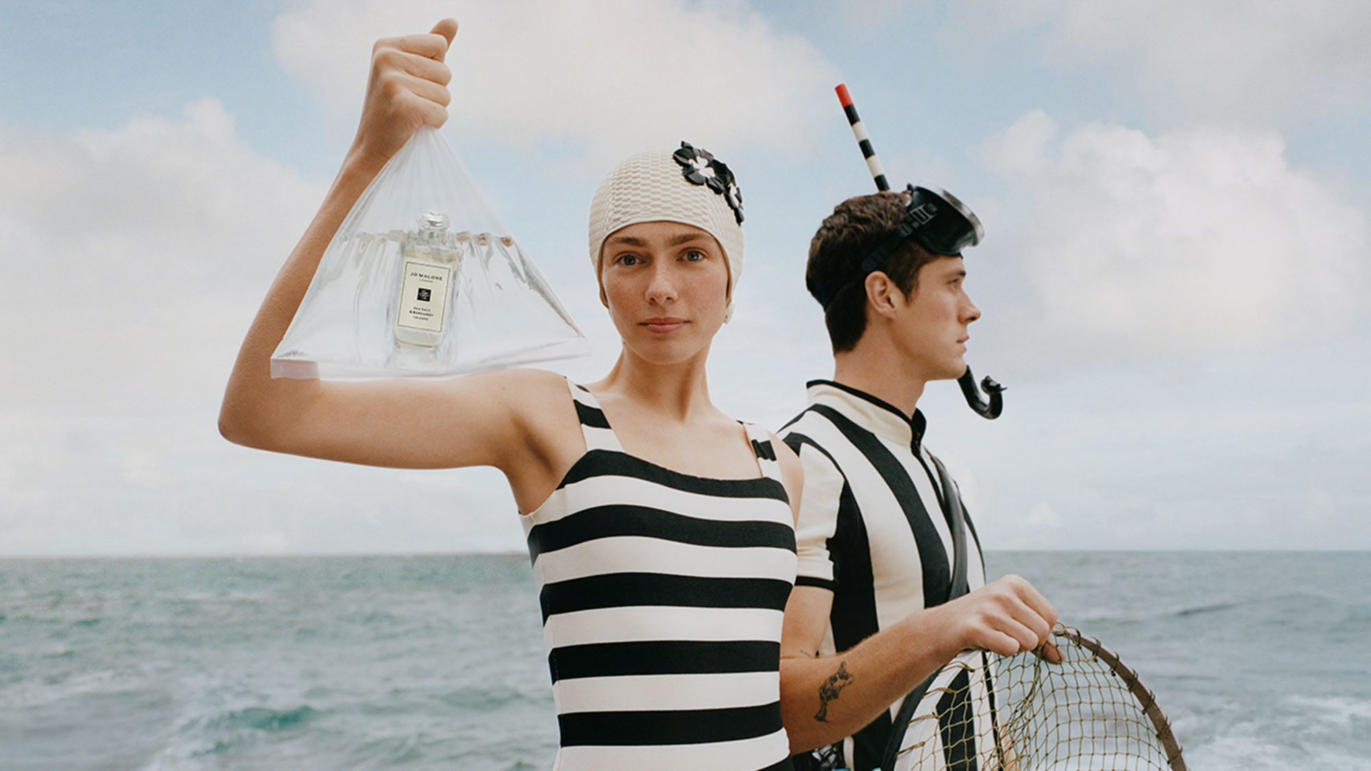 Two people in striped swimwear by the sea, one holding fragrance bottle in clear bag, wearing knit beanie and snorkel gear