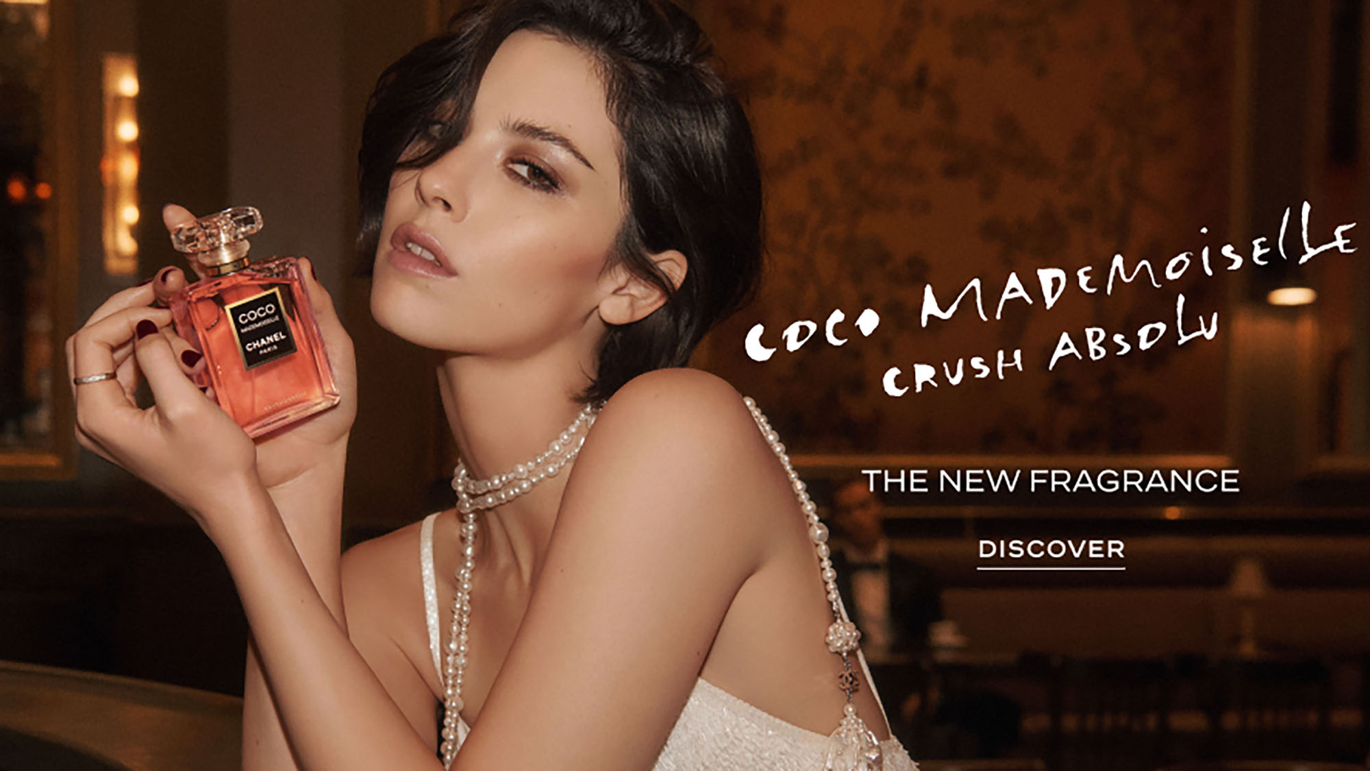 Woman holding coral fragrance bottle labelled Coco Mademoiselle Crush Absolu, wearing pearl jewellery against warm gold-toned background
