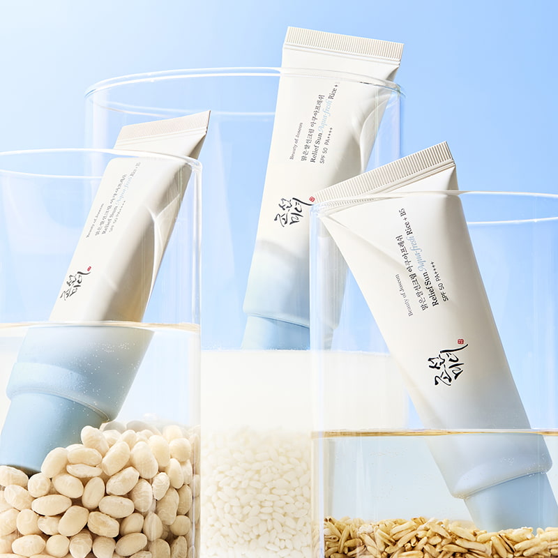 Three skincare tubes are displayed vertically in glass containers filled with different grains against a clear blue sky.