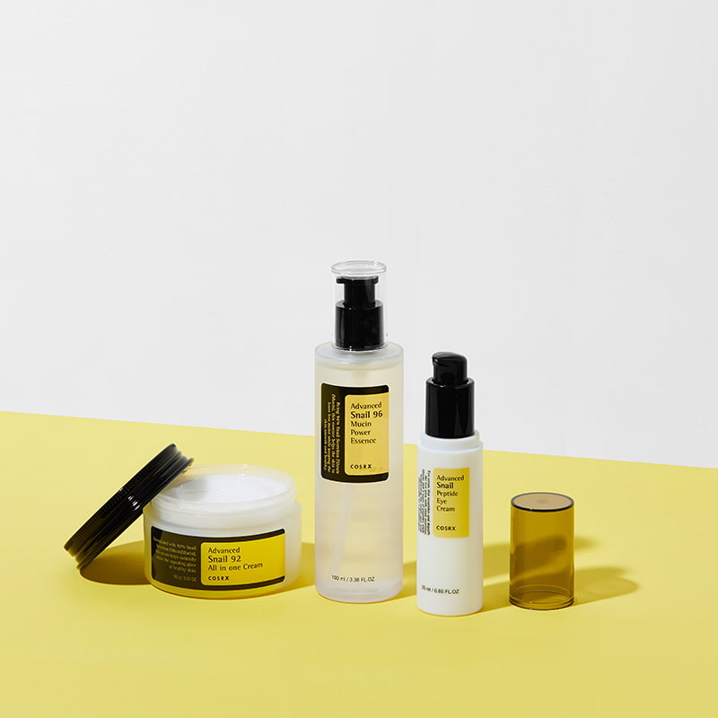 Skincare products with yellow labels, including a jar, two pump bottles, and a tube, arranged on a yellow and white background.