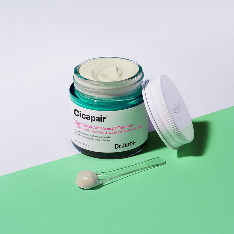 An open jar of Dr.Jart+ Cicapair Color Correcting Treatment with a small portion on a spatula, set against a green background.