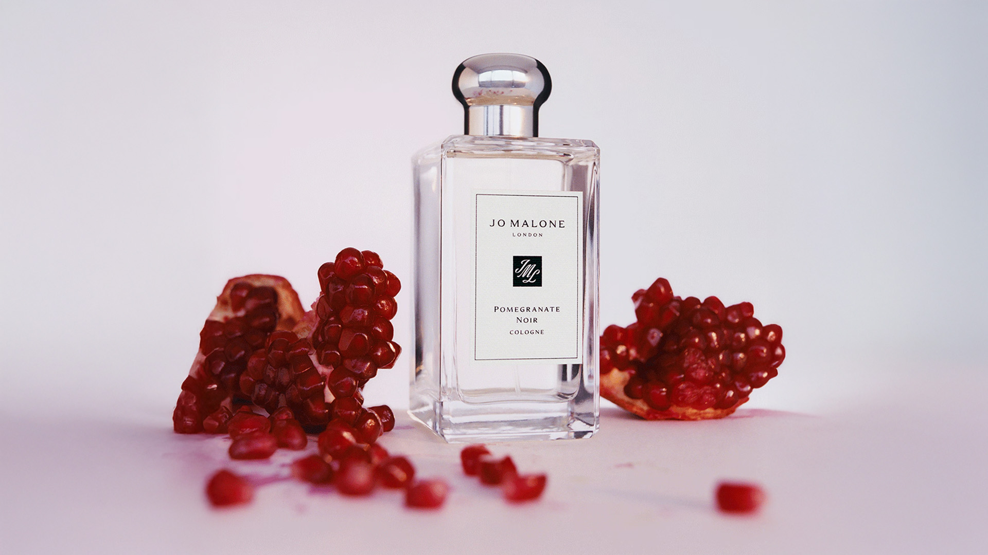 A bottle of Jo Malone cologne is placed between scattered pomegranate seeds and fruit pieces on a white surface.