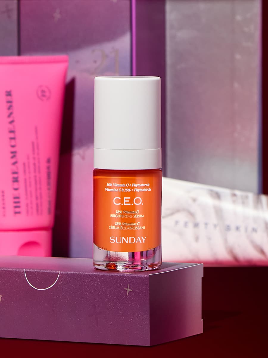 A bottle of C.E.O. Vitamin C serum with a white cap stands on a purple box, with other skincare products in the background.