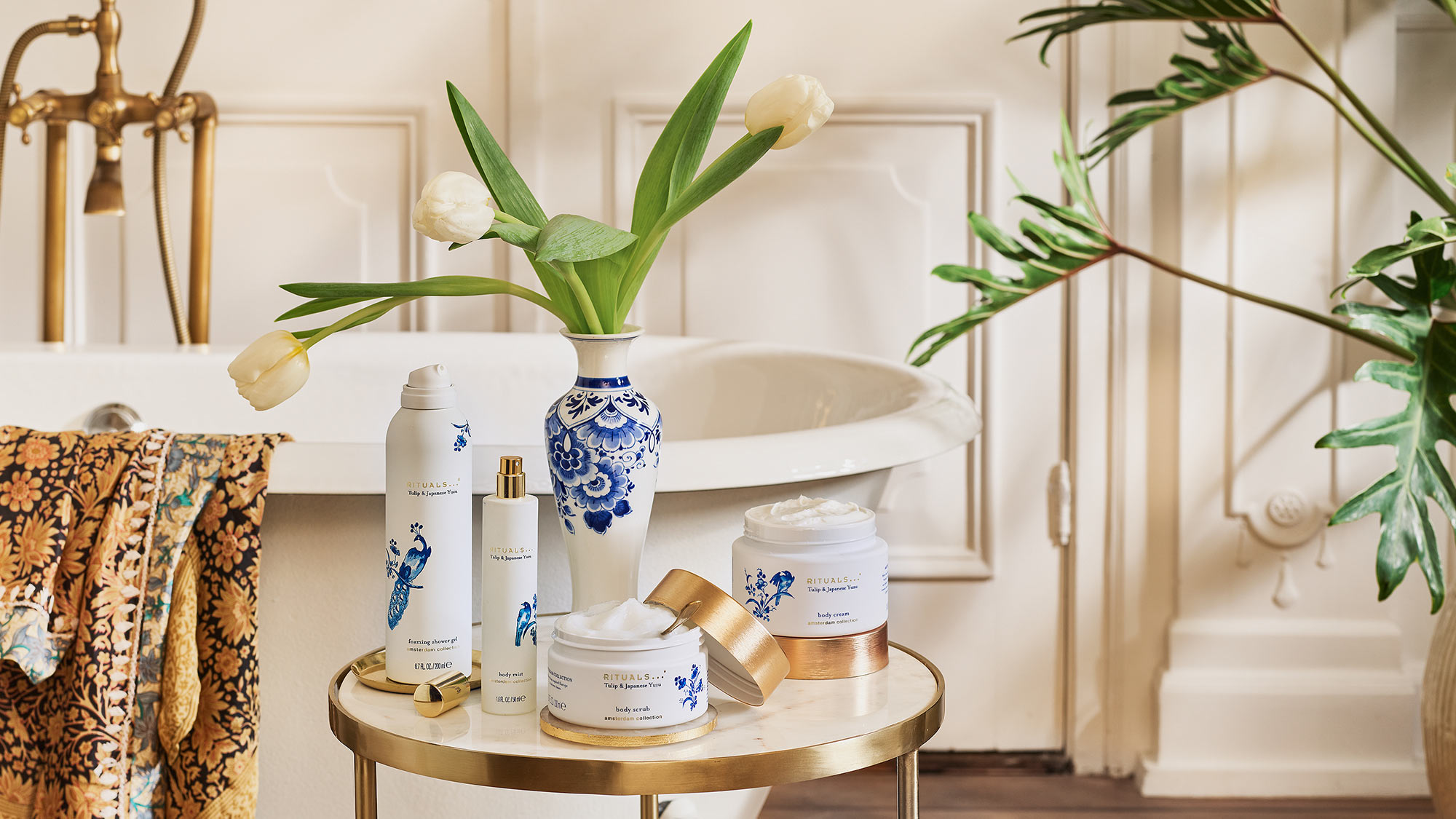 A white bathtub with a gold faucet is in a bathroom with a floral robe, a round table with skincare products, and a vase with tulips nearby.