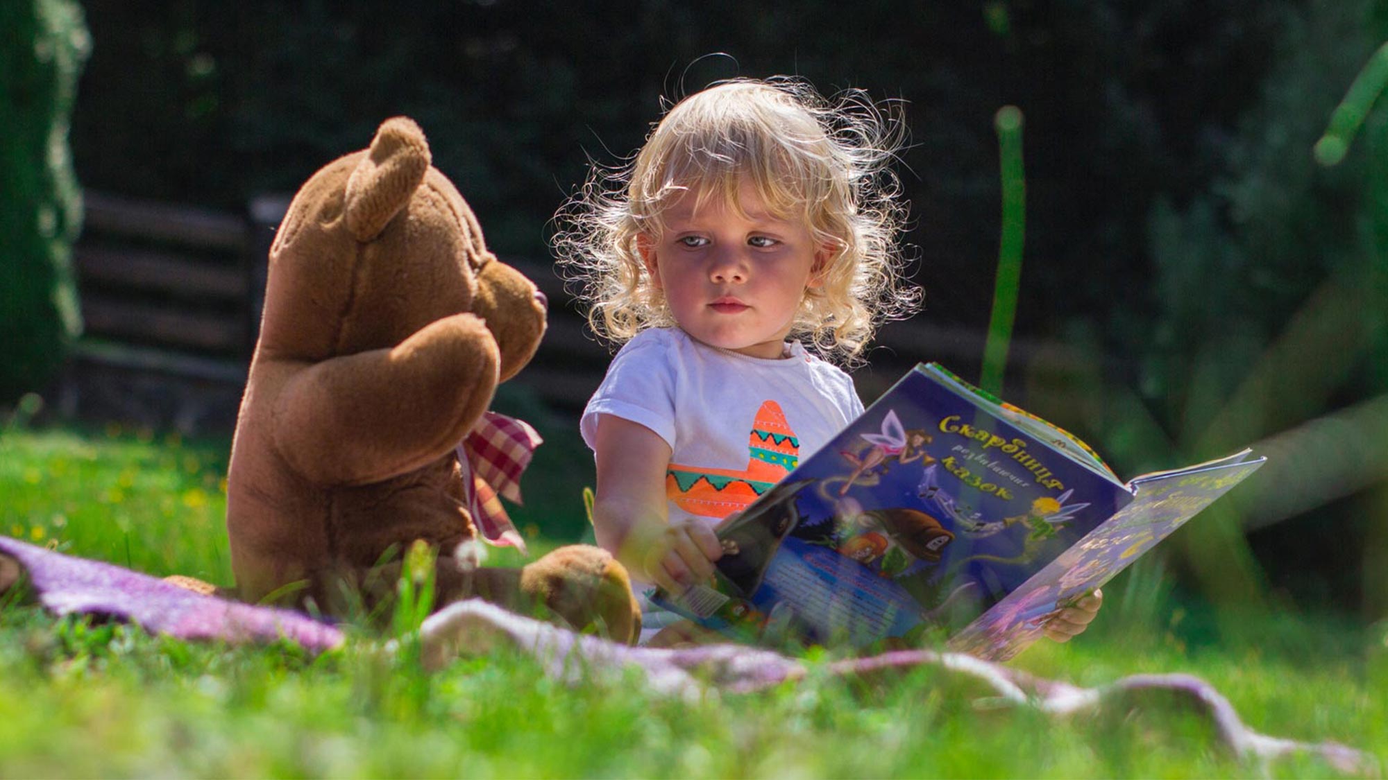 Young child with blonde curly hair reading a storybook whilst sitting outdoors with a brown teddy bear on grass and moss.