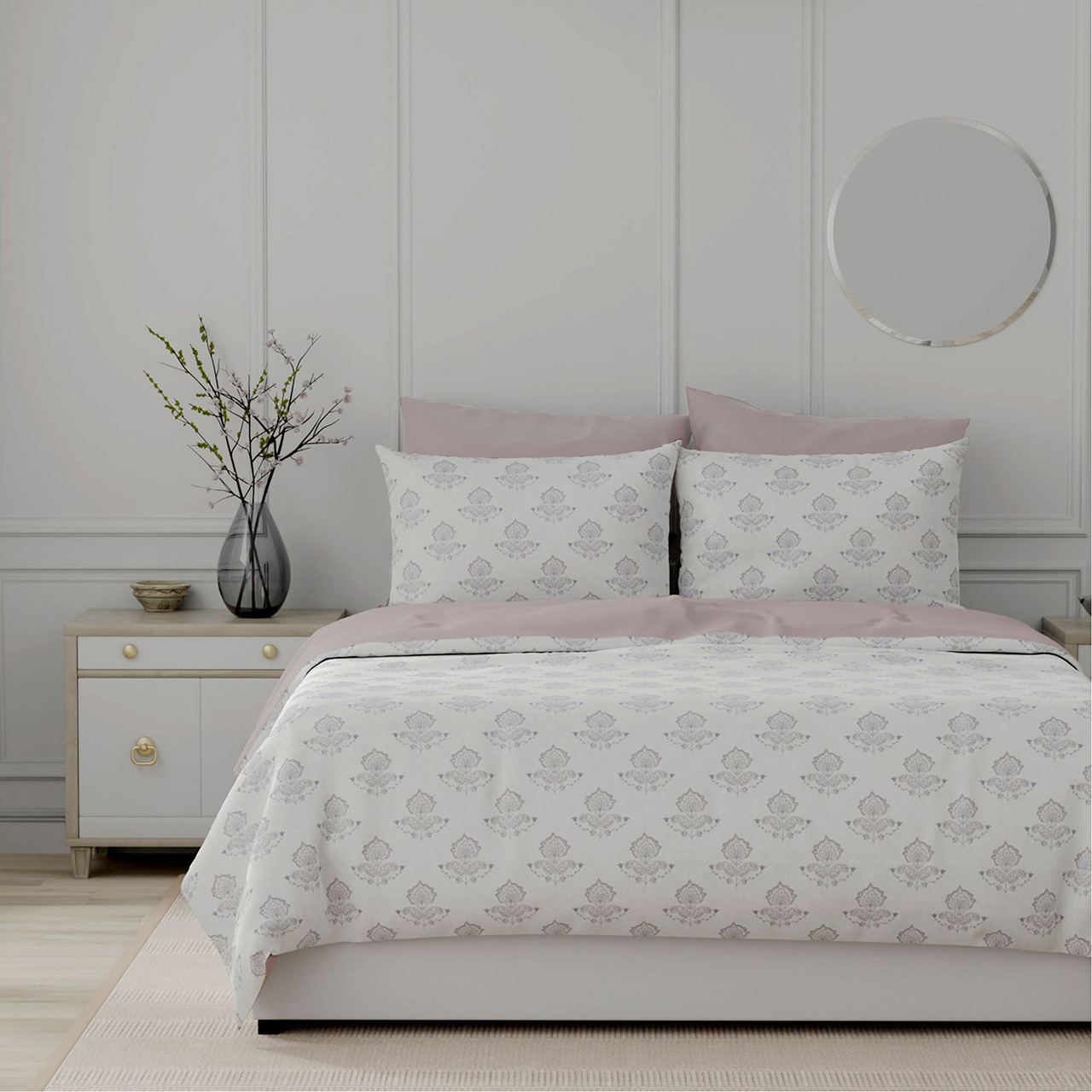Bedroom styled with patterned bedding in cream and pink tones, coordinating pillows, wooden bedside table with vase, and circular mirror on wall