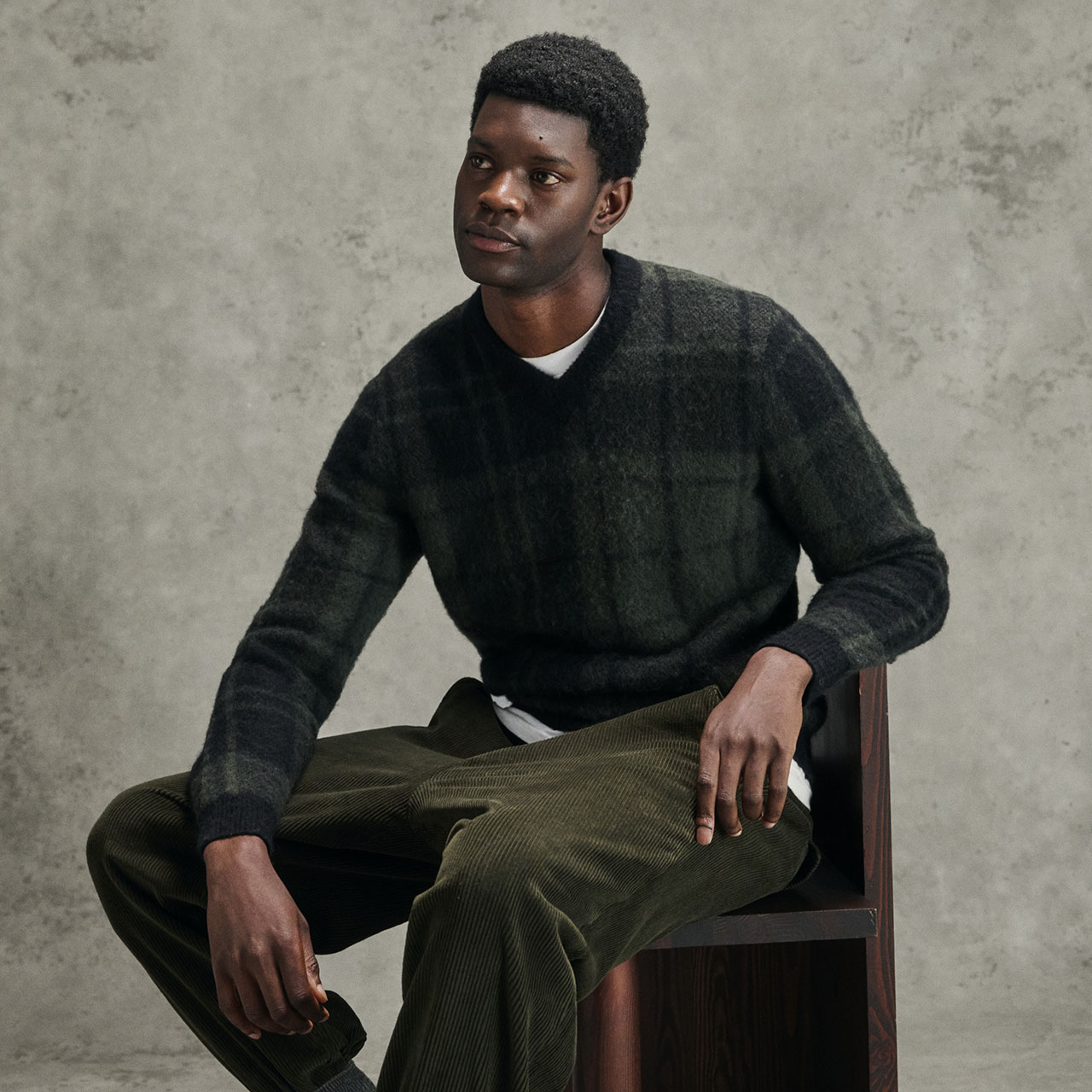 Man wearing charcoal knit jumper and olive corduroy trousers, seated against concrete background