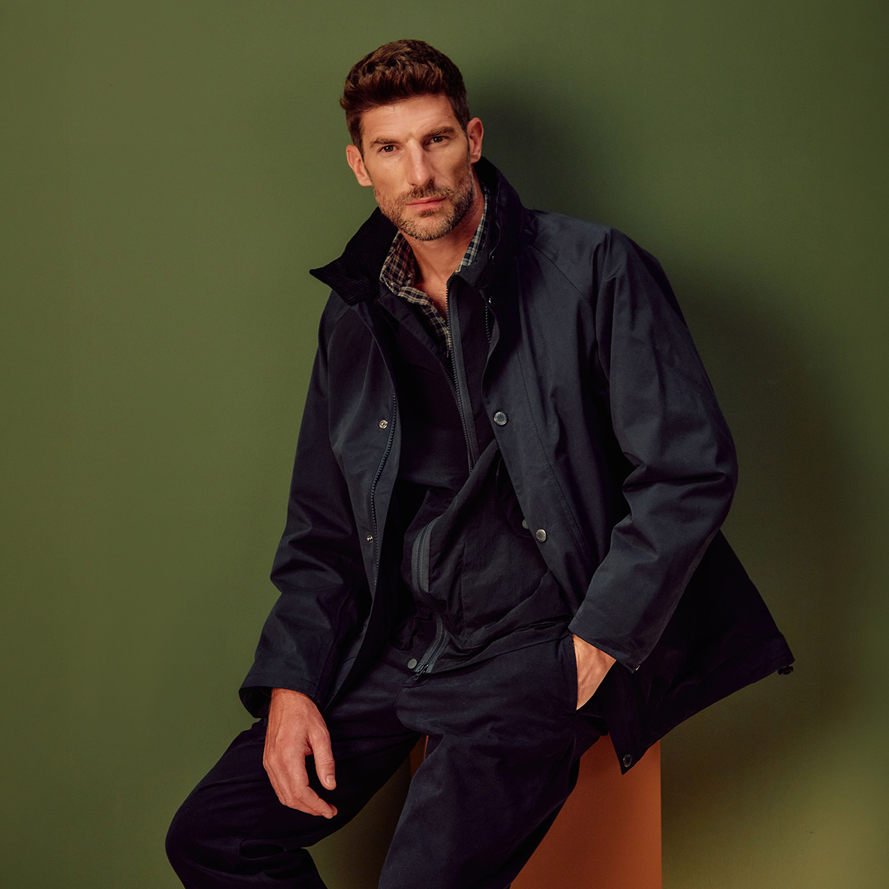 Man wearing navy jacket with checked shirt over navy trousers, sitting against green wall