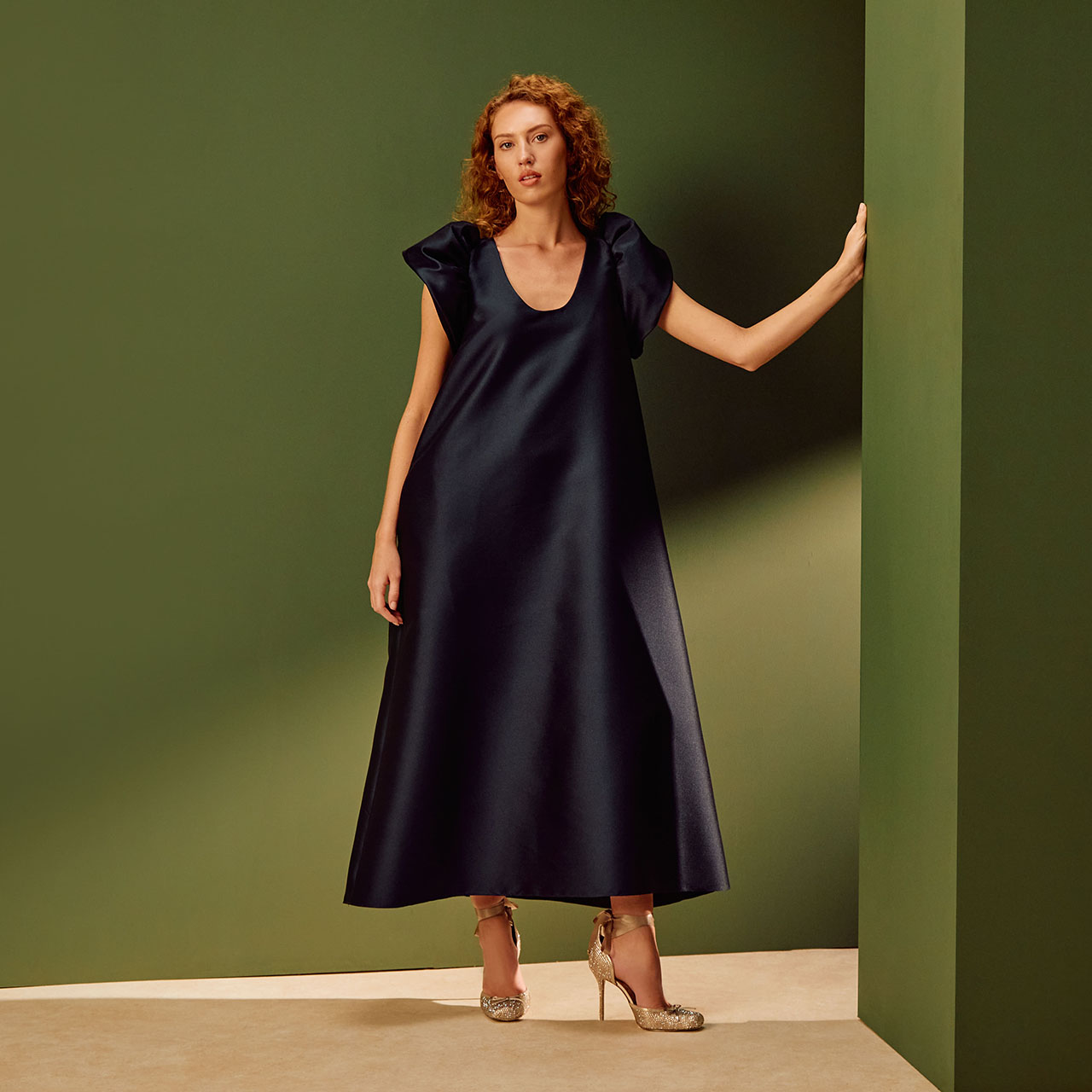 Woman with red curly hair wearing navy midi dress with cap sleeves and gold heeled sandals