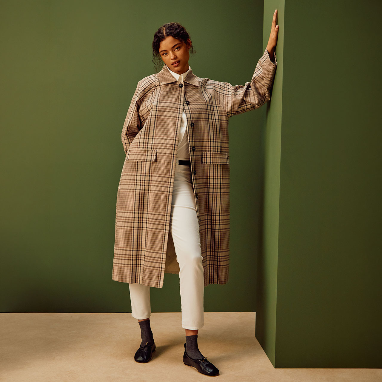 Woman wearing brown and cream checked coat over white trousers and black ankle boots against green wall
