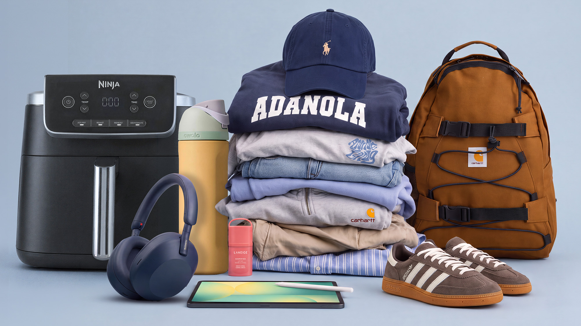 A collection of items including a Ninja air fryer, stack of folded clothes with a cap on top, brown backpack, black headphones, pale yellow bottle, small red item, tablet, and brown trainers with white stripes, all arranged on a light blue background.