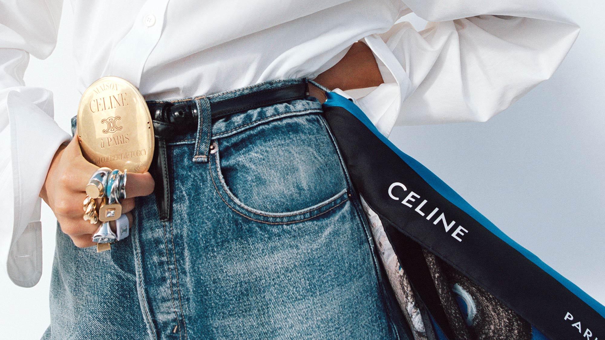 A woman in sunglasses, wearing a long dress, stands by the rocky shoreline holding a Celine bag.
