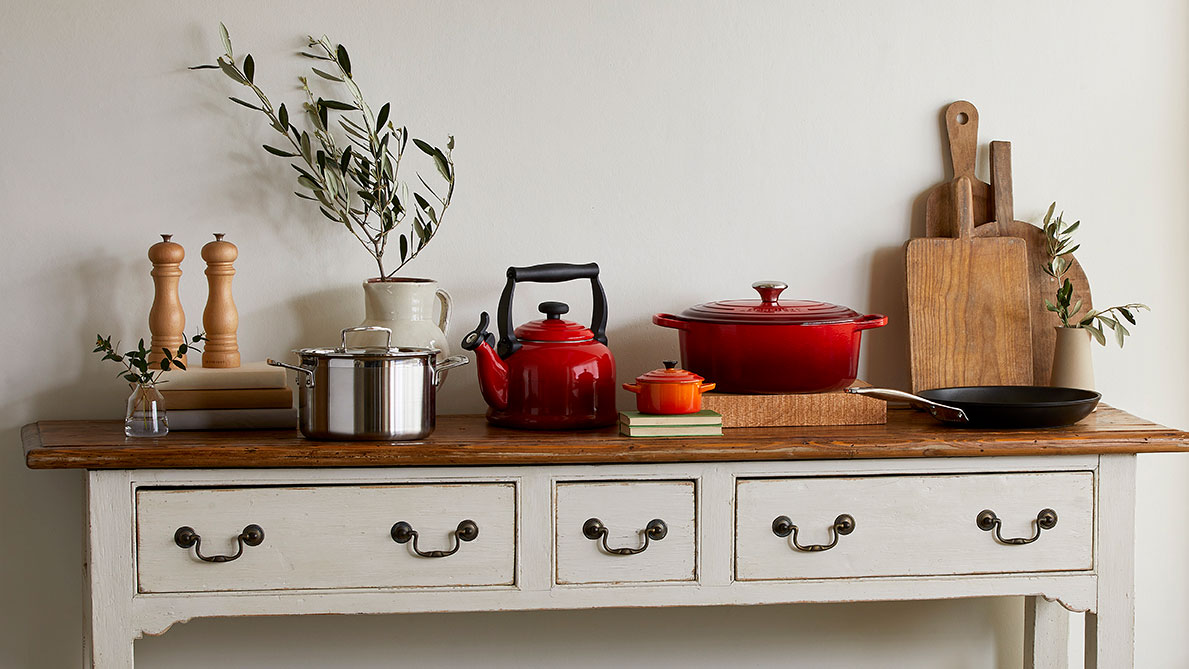 Kitchen worktop styled with red cookware, wooden chopping boards, stainless steel pot, and fresh herbs in vase