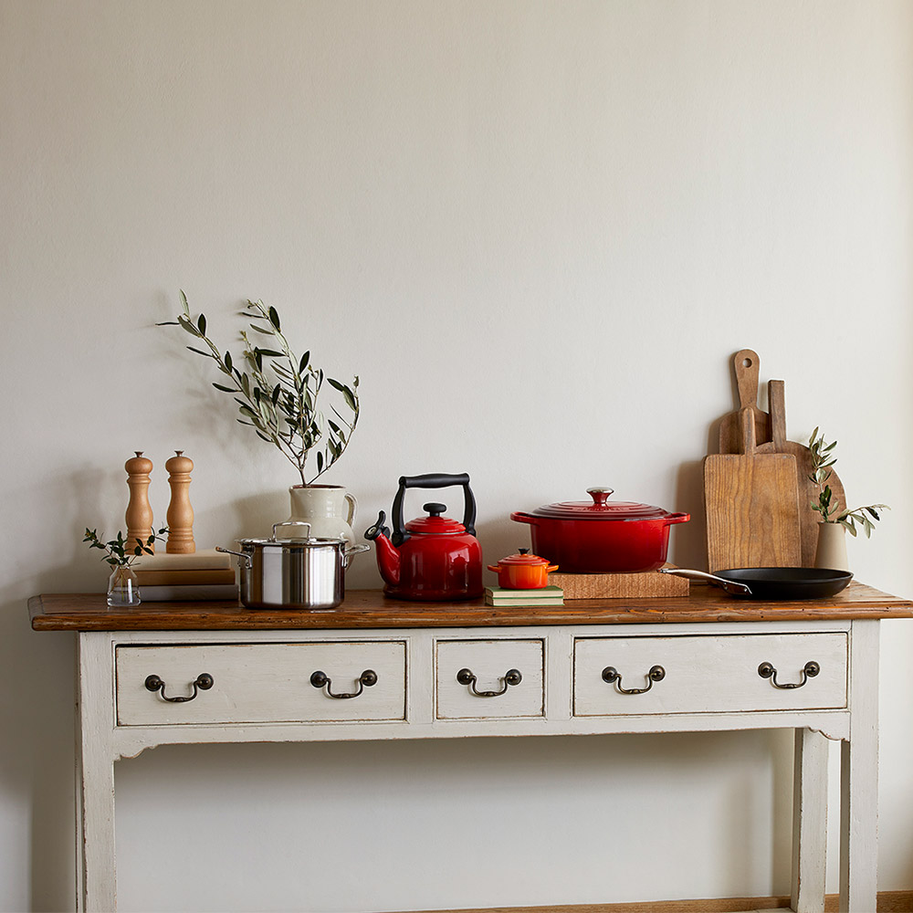 Kitchen worktop styled with red cookware, wooden chopping boards, stainless steel pot, and fresh herbs in vase