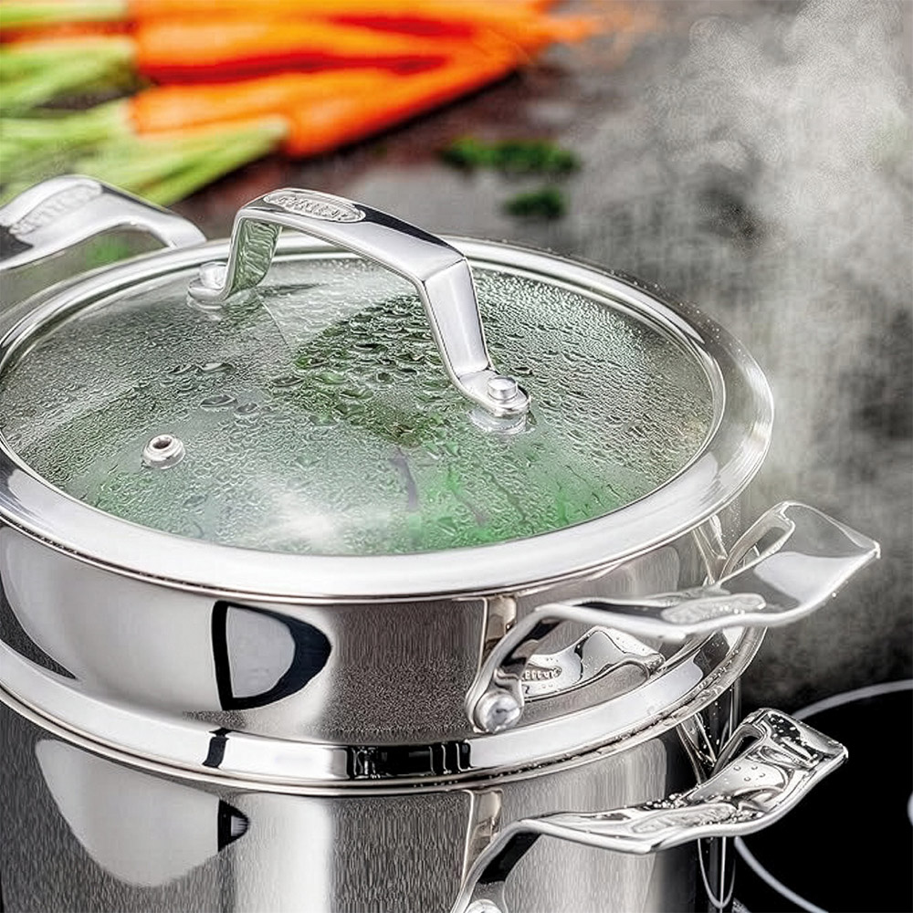 Stainless steel steamer pot with glass lid filled with steaming vegetables and fresh ingredients nearby