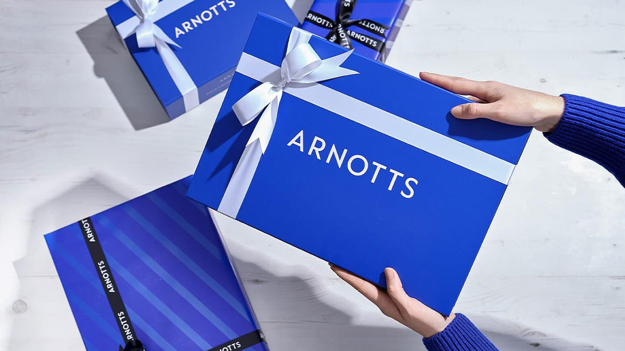 Hands holding a blue gift box with 'ARNOTTS' on it, surrounded by similar boxes with ribbons on a light surface.
