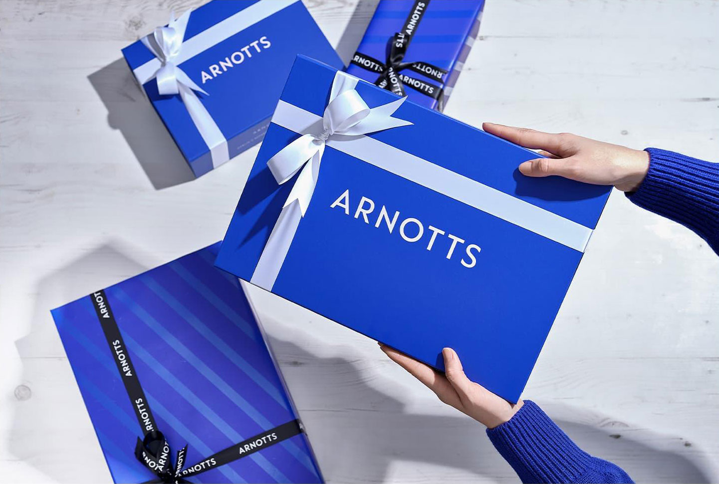 Hands holding a blue gift box with 'ARNOTTS' on it, surrounded by similar boxes with ribbons on a light surface.